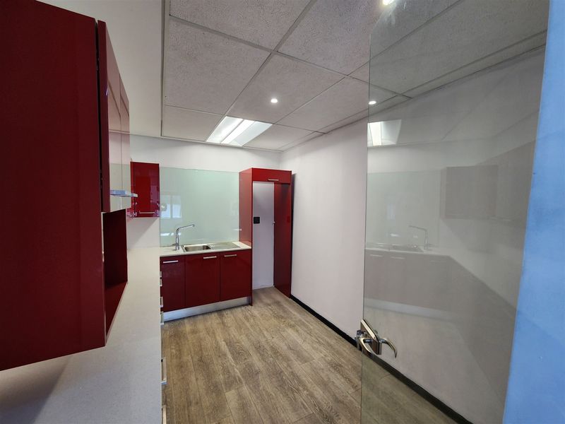 A modern office kitchenette featuring red high-gloss cabinetry, a stainless steel sink, and durable laminate flooring with a glass entry door.