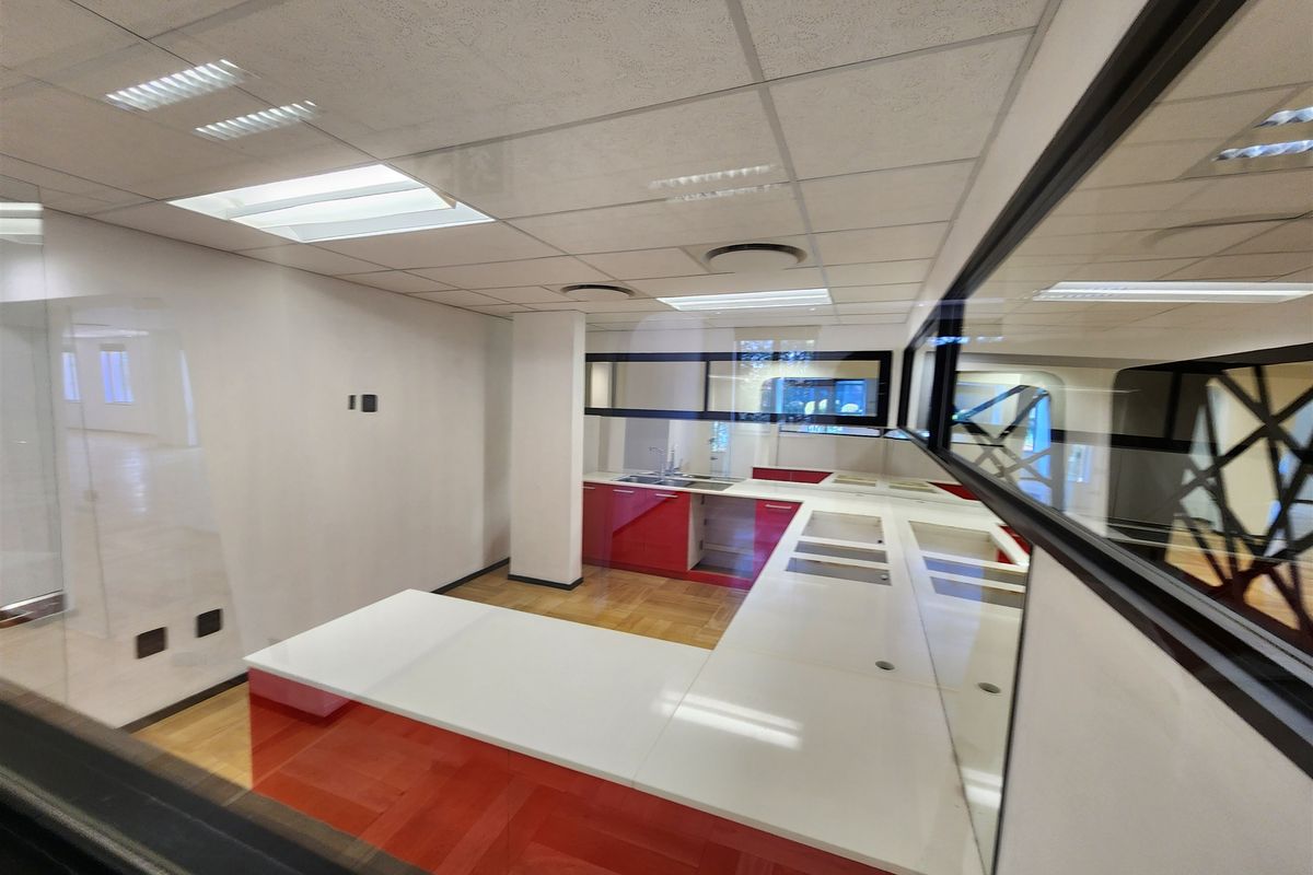 A vacant office kitchen area featuring red cabinetry, white countertops, and a glass partition wall, viewed through a window.