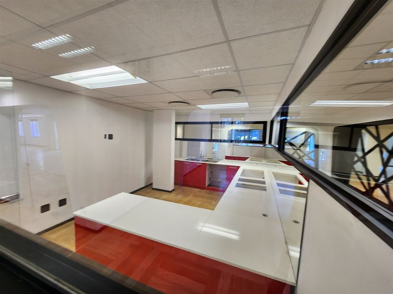A vacant office kitchen area featuring red cabinetry, white countertops, and a glass partition wall, viewed through a window.