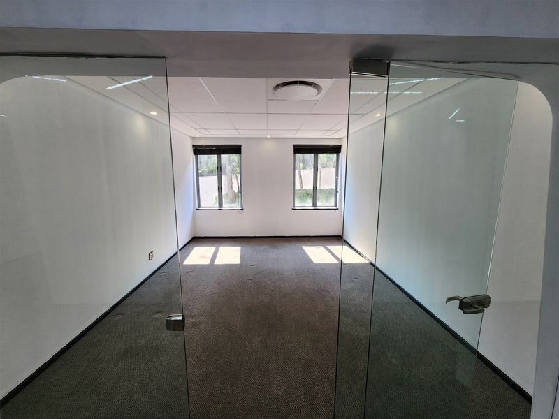 A bright, empty office space featuring glass partition doors, carpeted flooring, and a suspended ceiling with recessed lighting.