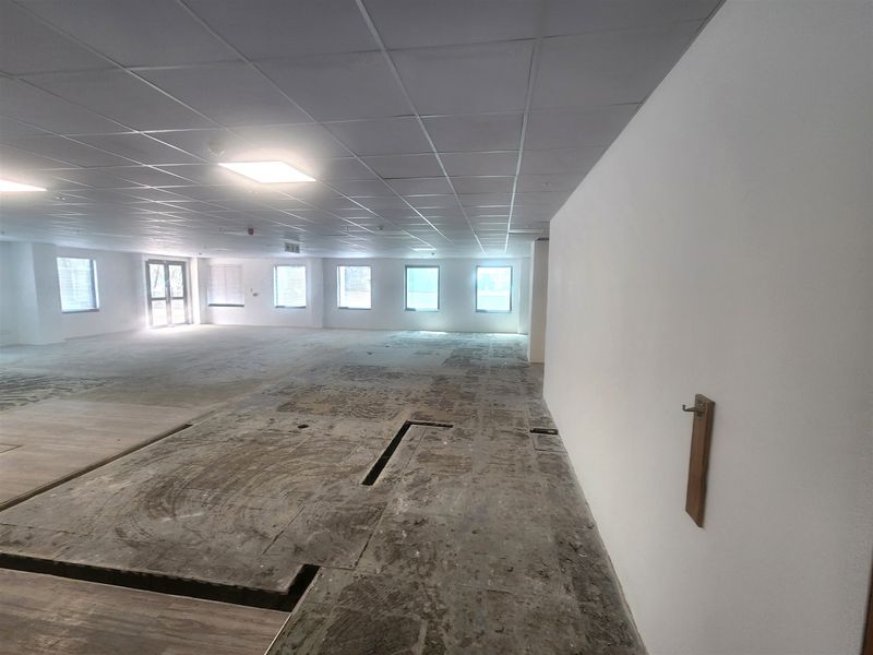A large, open-plan office space featuring a suspended ceiling, recessed lighting, and multiple windows, currently requiring flooring installation.