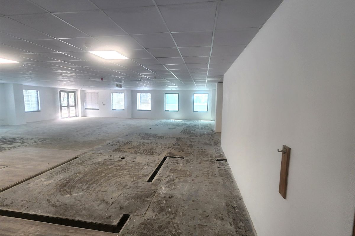 A large, open-plan office space featuring a suspended ceiling, recessed lighting, and multiple windows, currently requiring flooring installation.