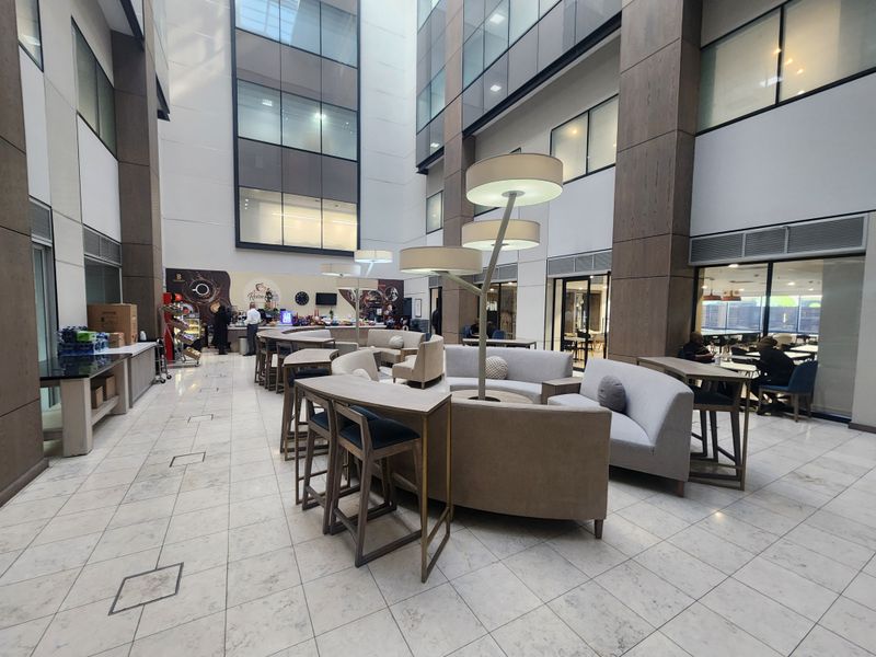 A modern, high-ceilinged open-plan office atrium featuring marble flooring, expansive glass facades, and a central integrated coffee bar area.
