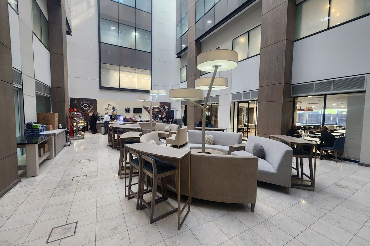 A modern, high-ceilinged open-plan office atrium featuring marble flooring, expansive glass facades, and a central integrated coffee bar area.