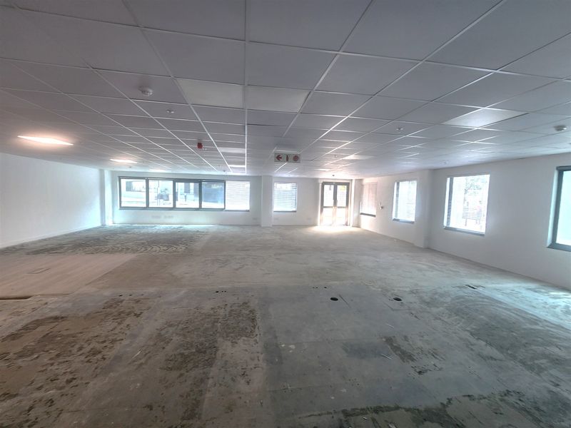 A spacious, open-plan office area featuring a suspended ceiling, recessed lighting, and large windows with blinds, currently awaiting final flooring installation.
