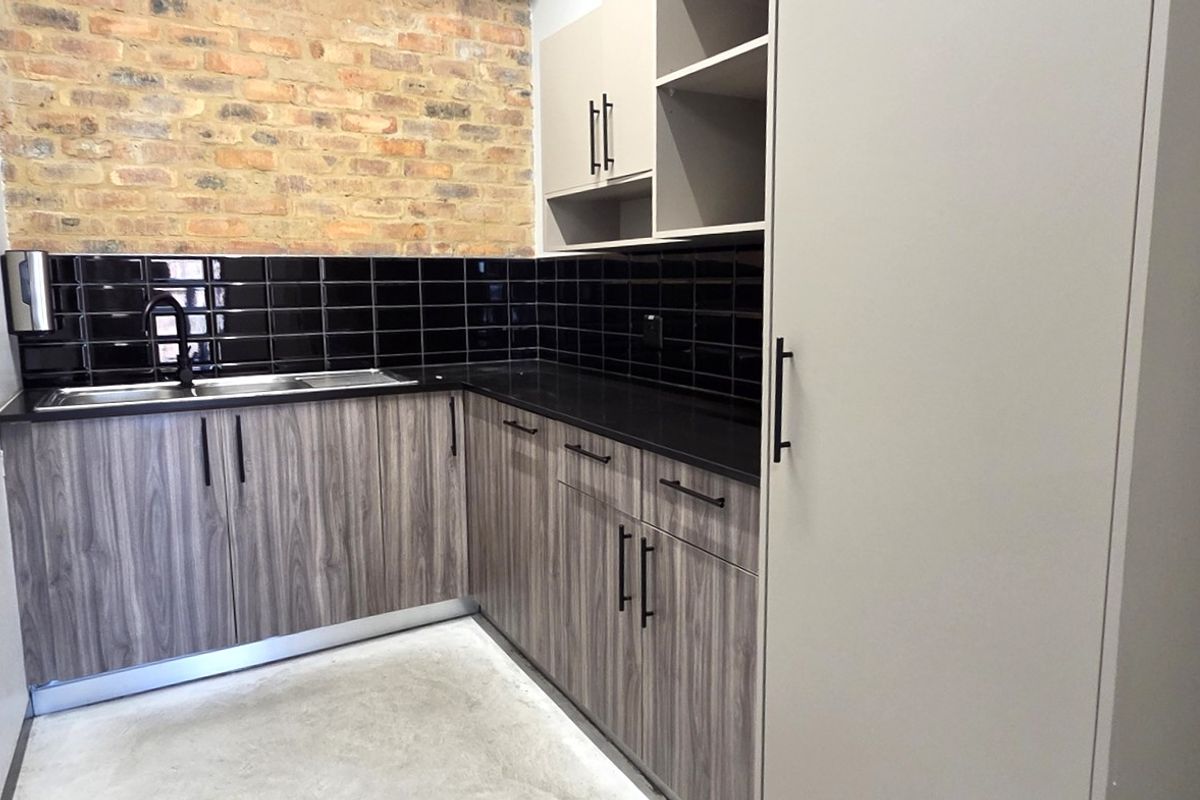 A modern kitchenette featuring wood-grain cabinetry, black subway tile backsplash, exposed brick accent wall, and a stainless steel sink with a black faucet.