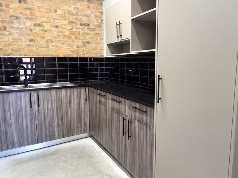 A modern kitchenette featuring wood-grain cabinetry, black subway tile backsplash, exposed brick accent wall, and a stainless steel sink with a black faucet.