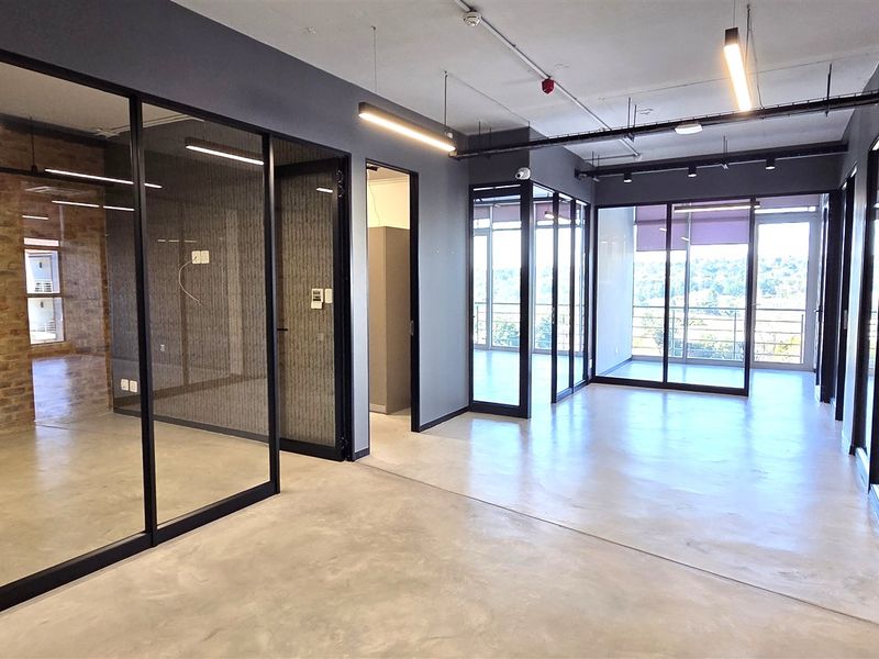 Modern open-plan office space featuring polished concrete floors, industrial-style exposed ceilings, glass partitions, and an exposed brick feature wall with balcony access.