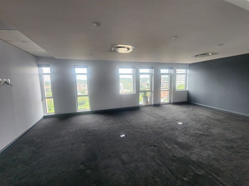 A spacious, empty office area featuring dark carpeting, recessed lighting, and large windows providing ample natural light and exterior views.