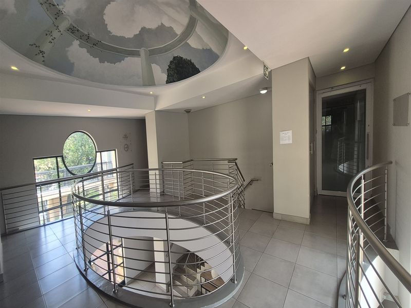 A bright office building hallway featuring a central spiral staircase with stainless steel balustrades, tiled flooring, and a decorative circular ceiling mural.
