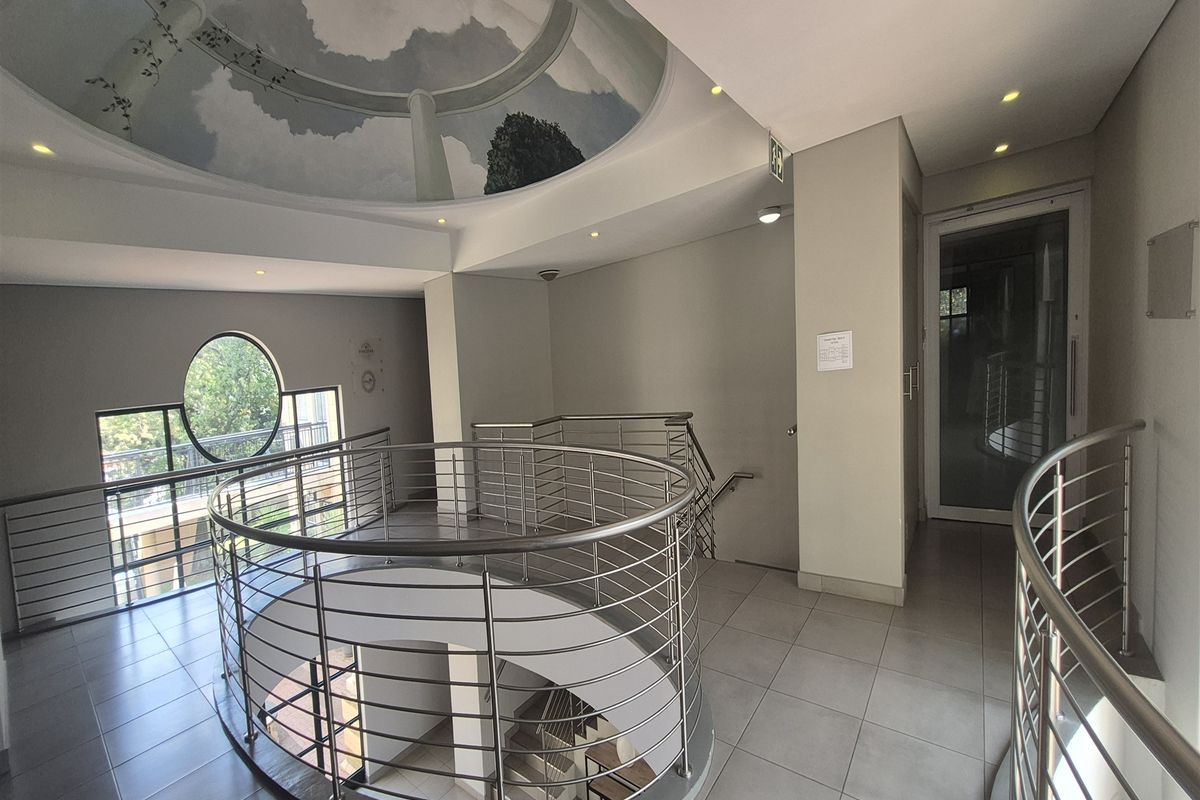A bright office building hallway featuring a central spiral staircase with stainless steel balustrades, tiled flooring, and a decorative circular ceiling mural.