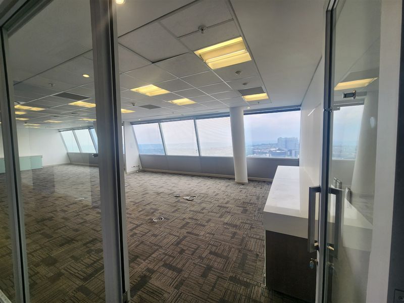 A spacious, empty office suite featuring floor-to-ceiling windows with city views, suspended ceilings, and carpeted flooring, viewed through a glass partition entrance.