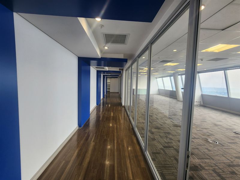 A modern office hallway featuring polished hardwood floors, blue accent pillars, and glass partitions looking into an open-plan workspace with carpeted flooring.