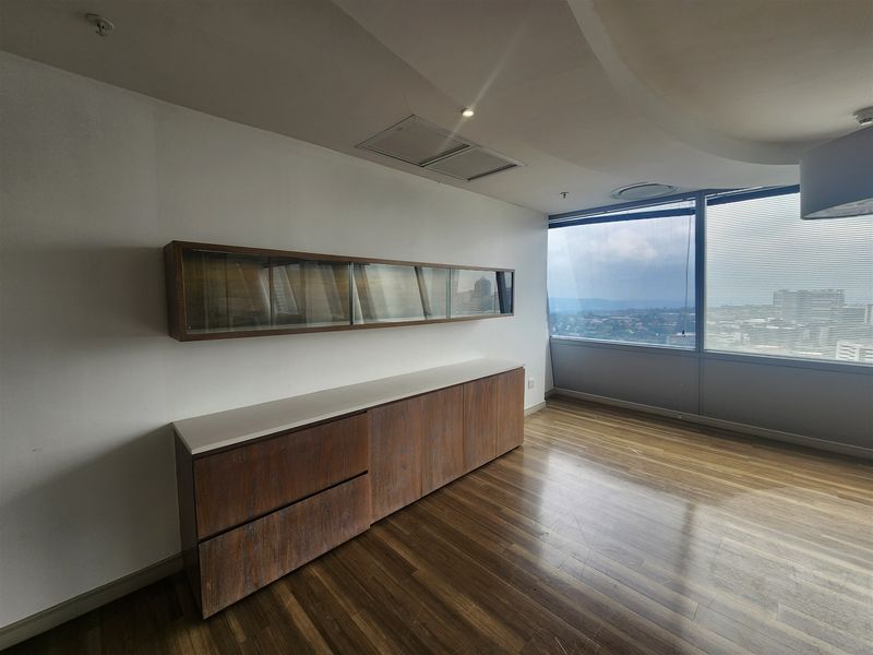 A bright office space featuring built-in wooden cabinetry, wall-mounted storage, hardwood flooring, and expansive windows offering panoramic city views.