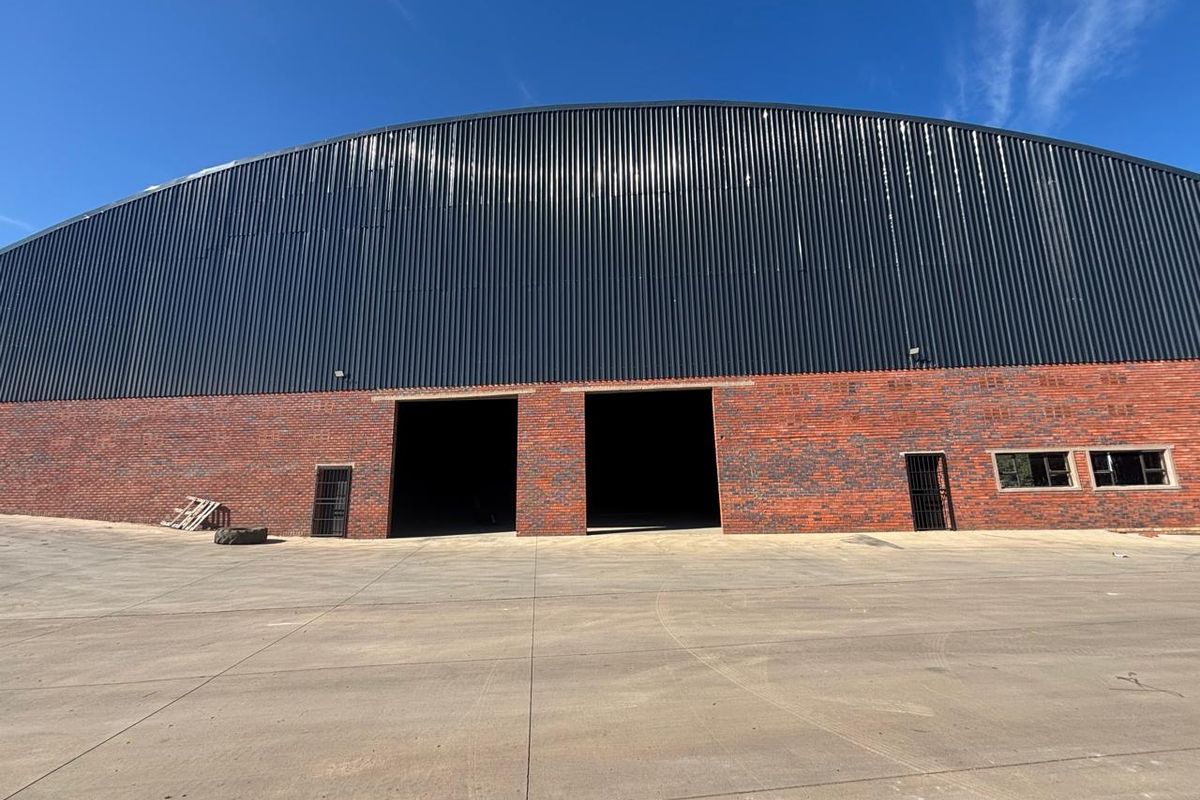 Industrial warehouse exterior featuring a large corrugated metal roof, brick facade, and expansive concrete yard with dual industrial access points.