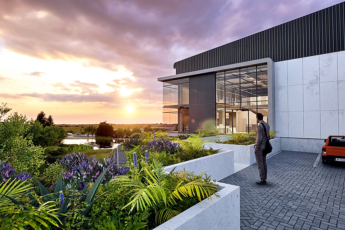 Modern industrial facility exterior featuring a large glass facade, concrete landscaping planters, and a paved driveway at sunset.