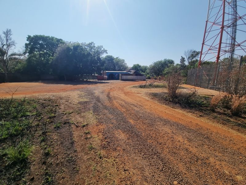 A wide-angle view of the property entrance featuring a gravel driveway, security fencing, and a prominent telecommunications tower on the site.