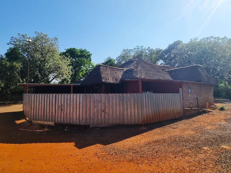 Exterior view of a farm-style dwelling featuring a traditional thatch roof and corrugated metal perimeter fencing on a dirt access path.