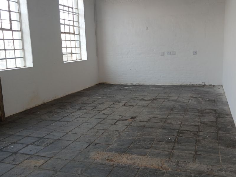 An empty industrial-style office space featuring exposed brick walls, large steel-framed windows, and original stone tile flooring, offering a blank canvas for renovation.