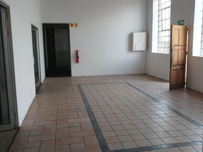 An empty industrial office space featuring tiled flooring, exposed ceiling beams, and large steel-framed windows providing ample natural light.