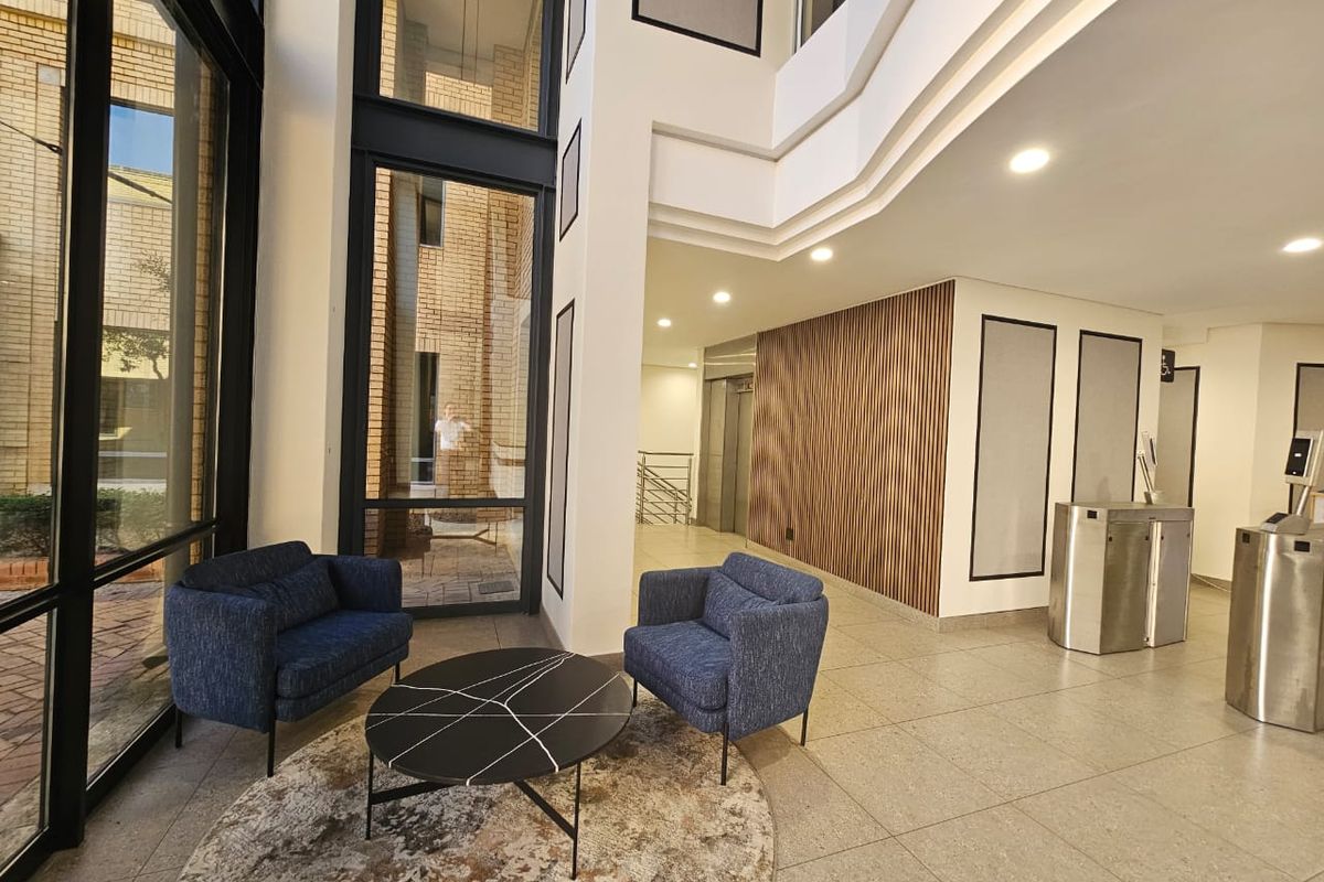Modern office building entrance lobby featuring double-volume windows, timber slat wall accents, security turnstiles, and elevator access.
