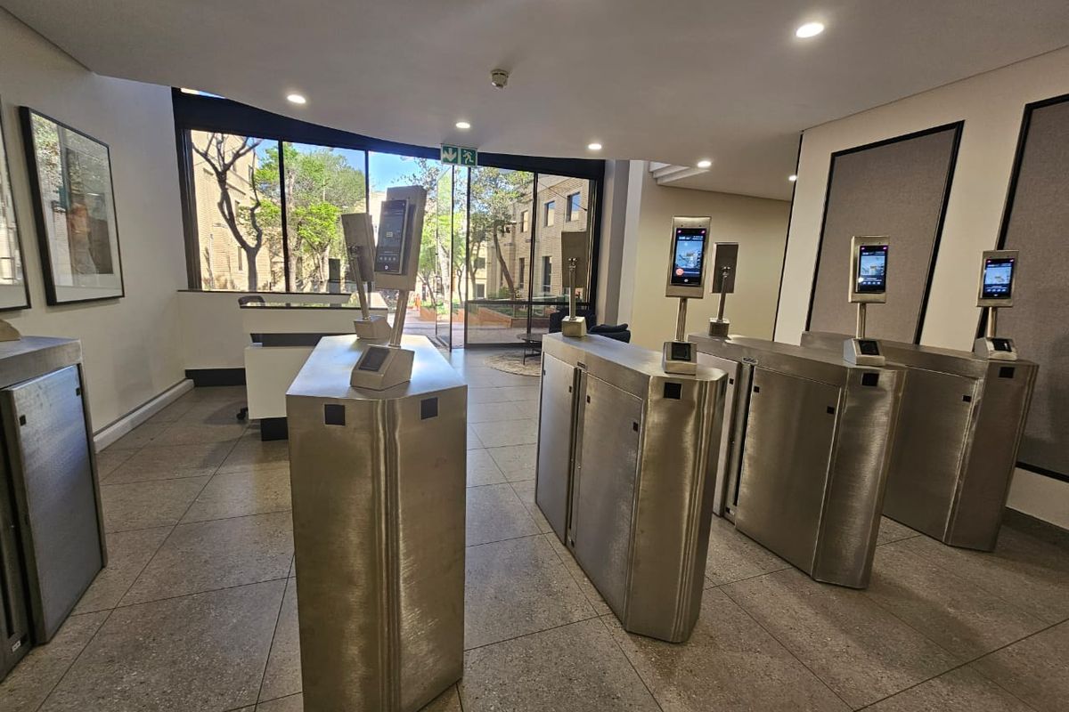Modern office building entrance lobby featuring stainless steel biometric access control turnstiles and large glass windows overlooking the exterior courtyard.