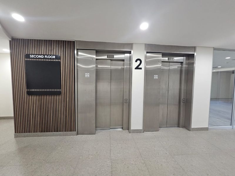 Modern office building elevator lobby on the second floor featuring stainless steel lift doors, slatted timber wall accents, and clean terrazzo flooring.