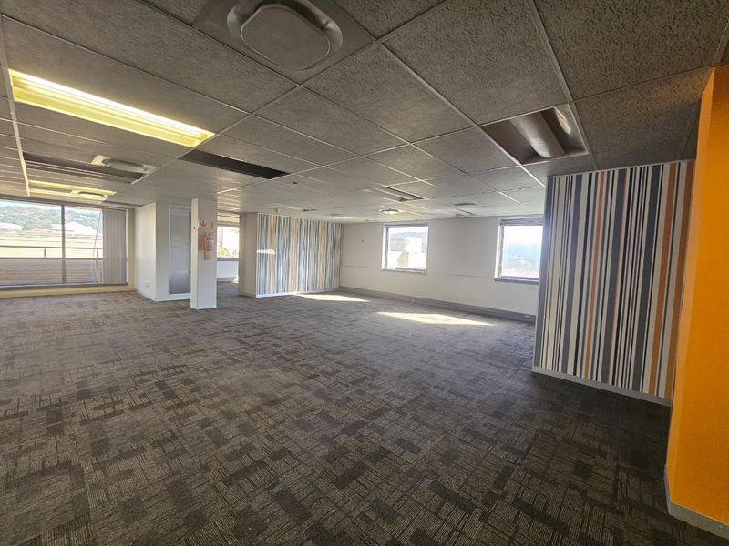 An open-plan office space featuring carpet tiles, a suspended ceiling with recessed lighting, and large windows providing natural light.