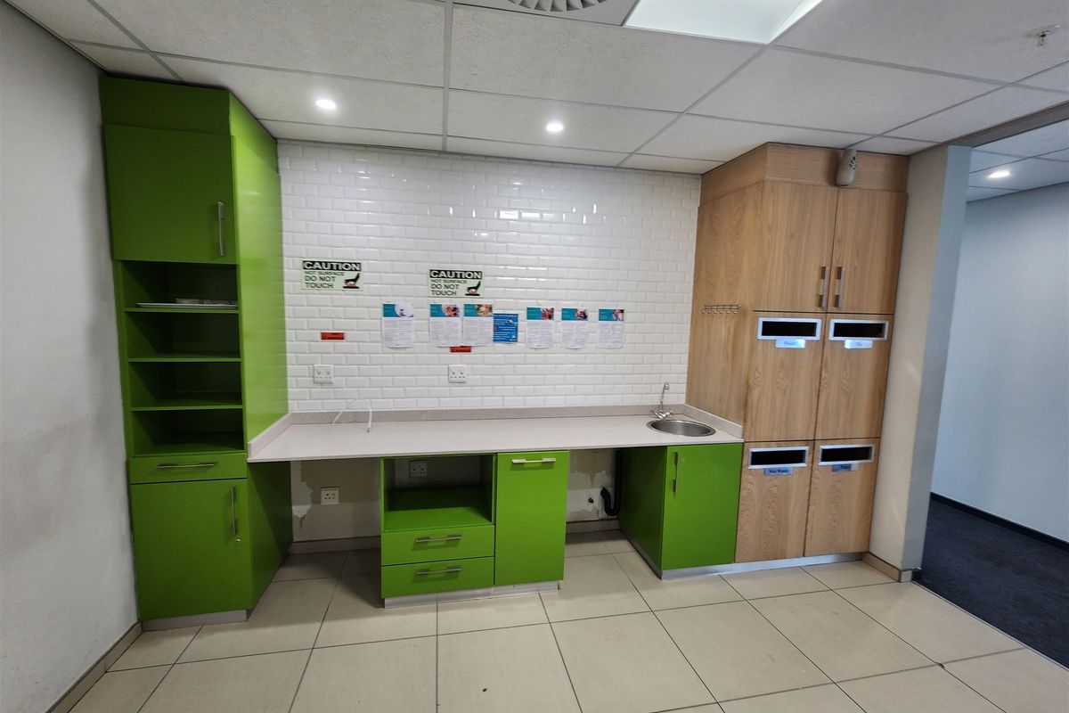 A functional office kitchenette featuring green and wood-grain cabinetry, white subway tile backsplash, integrated sink, and tiled flooring.