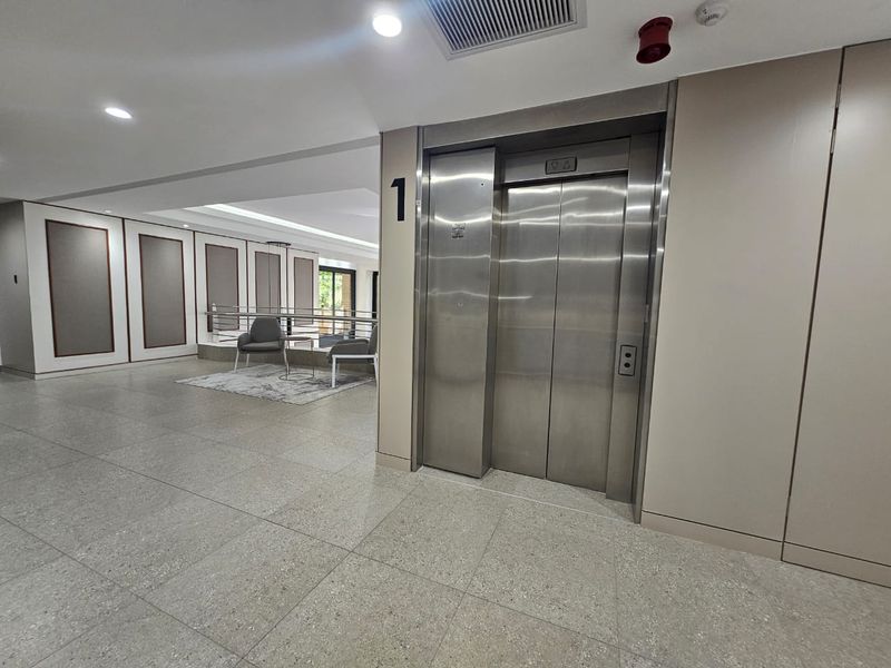 Modern office building hallway featuring a stainless steel elevator, recessed lighting, and clean tiled flooring with decorative wall paneling.