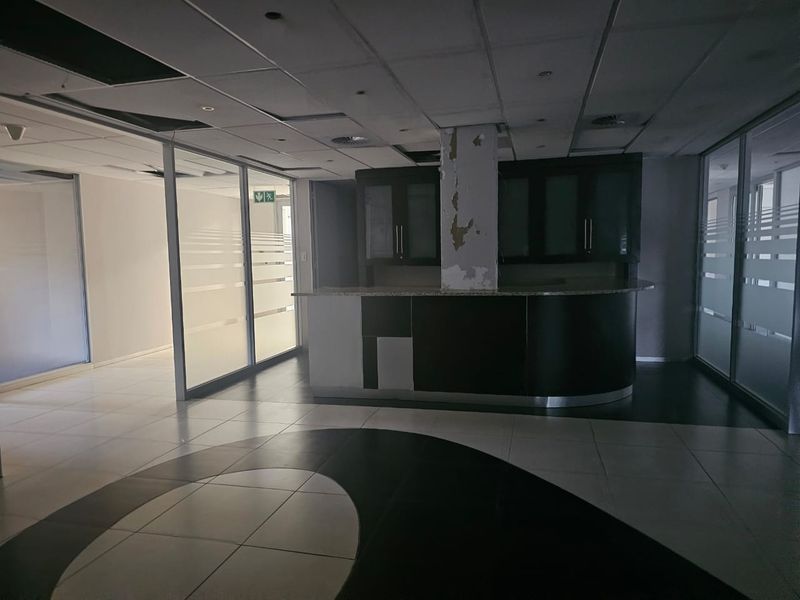 Empty office space featuring glass partitions, a built-in kitchenette counter, and tiled flooring, currently requiring maintenance to the ceiling and central pillar.