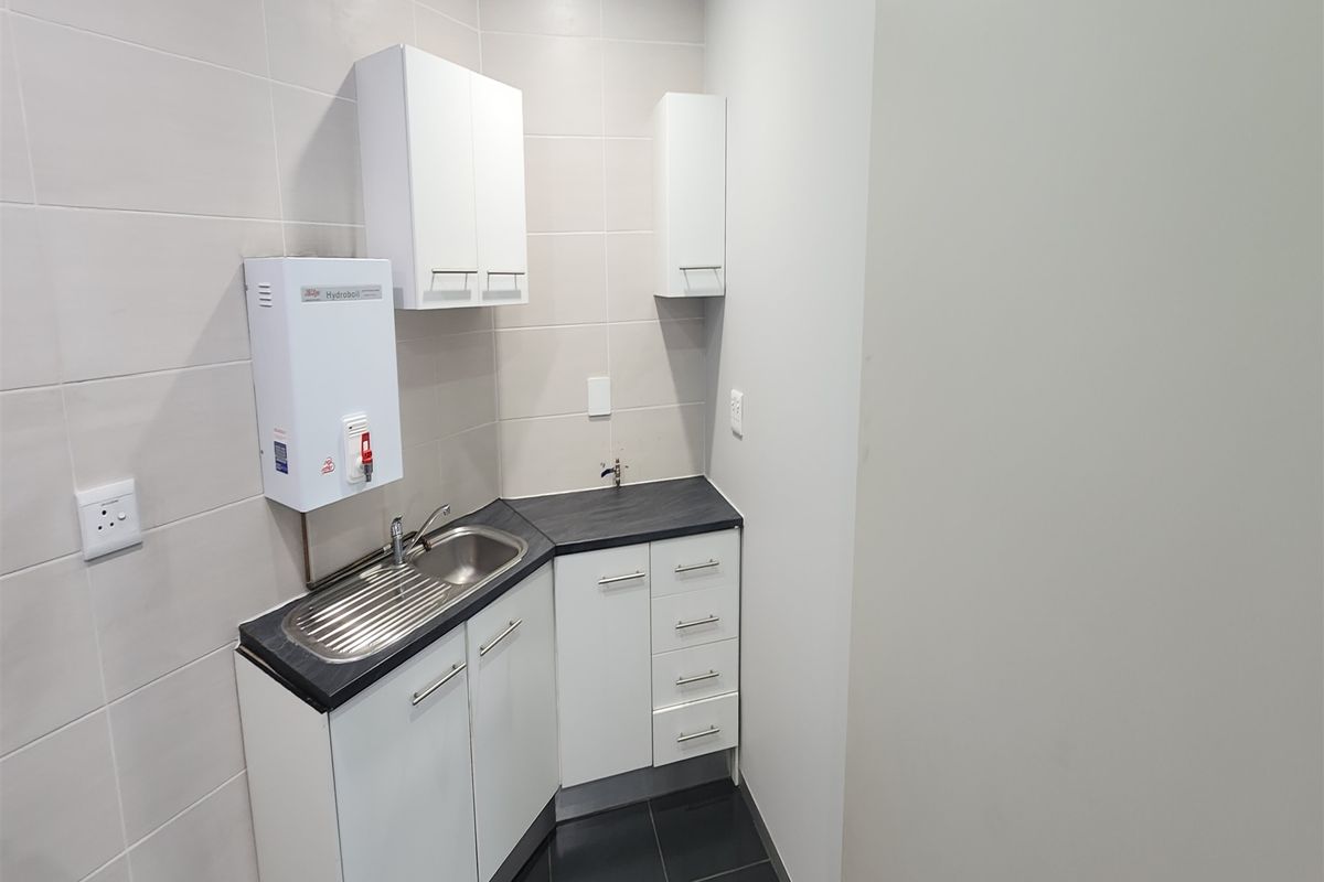 A compact office kitchenette featuring white cabinetry, a dark countertop, stainless steel sink, and a wall-mounted water boiler.