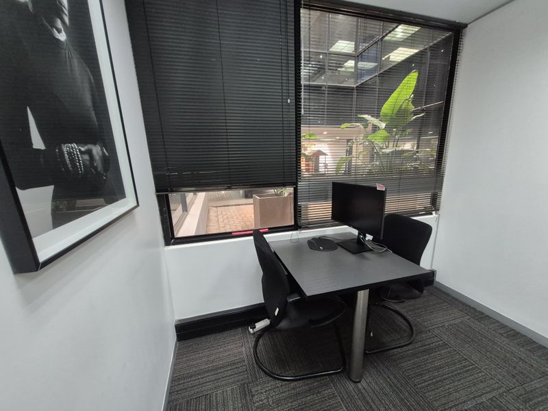 A compact office space featuring carpeted flooring, large windows with venetian blinds, and a view into an internal courtyard area.