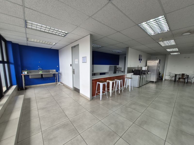Open-plan office break area featuring a kitchenette with a breakfast bar, stainless steel sink, tiled flooring, and suspended ceiling lighting.