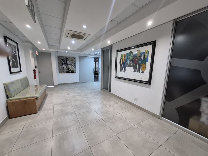 A modern office hallway featuring tiled flooring, recessed ceiling lighting, a ducted air conditioning unit, and glass partition doors.