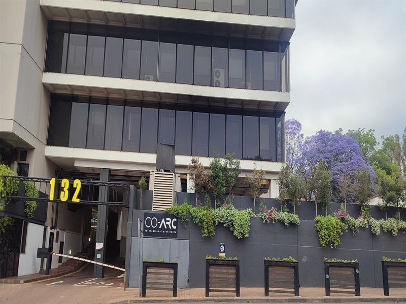 Front elevation of a multi-story office building featuring a modern tinted glass facade, secure gated entrance, and landscaped retaining wall.