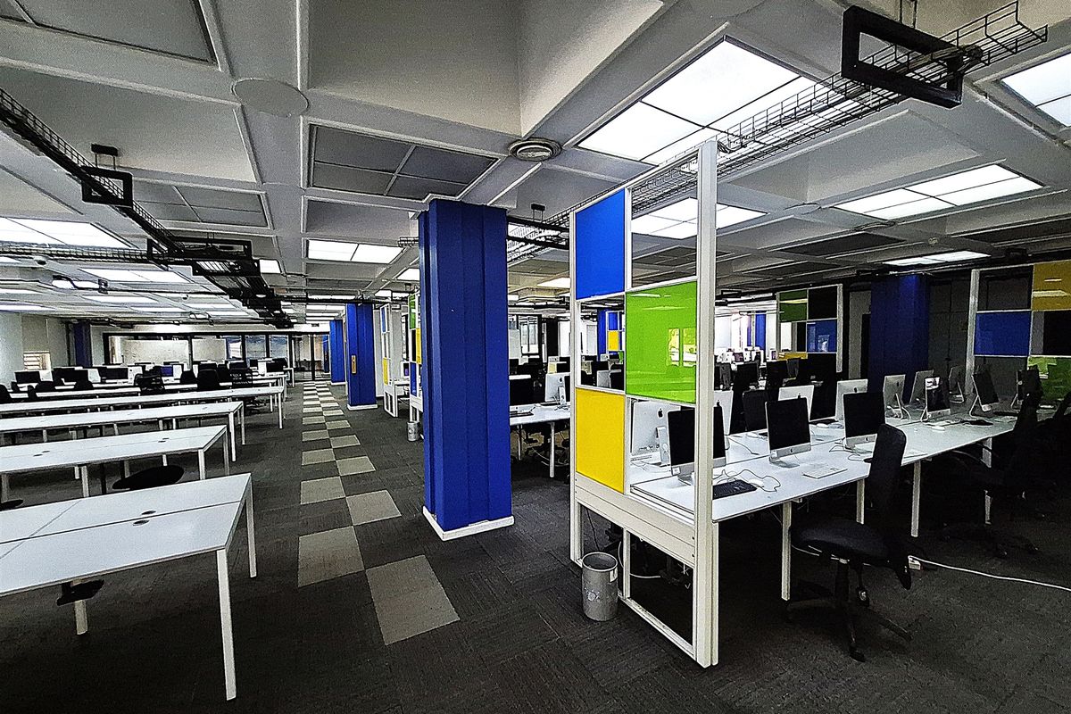 Open-plan office space featuring a suspended ceiling with recessed lighting, exposed cable management, and integrated desk partitions with a carpeted floor.