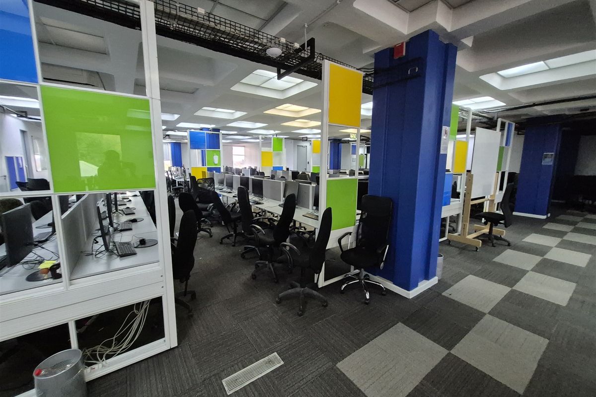 Open-plan office space featuring modular workstations, colorful partition screens, suspended ceiling grid, and carpeted flooring with integrated cable management systems.