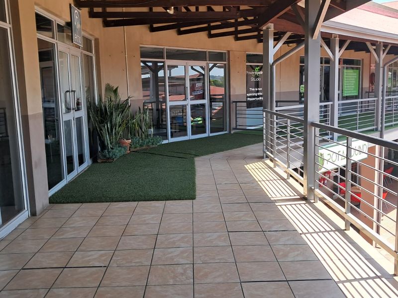 A covered outdoor walkway or balcony area featuring tiled flooring, a metal safety railing, and glass-fronted office entrances.