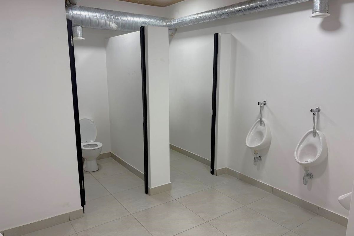 A clean, modern industrial-style communal restroom featuring tiled flooring, wall-mounted urinals, and private toilet cubicles with exposed ceiling ducting.