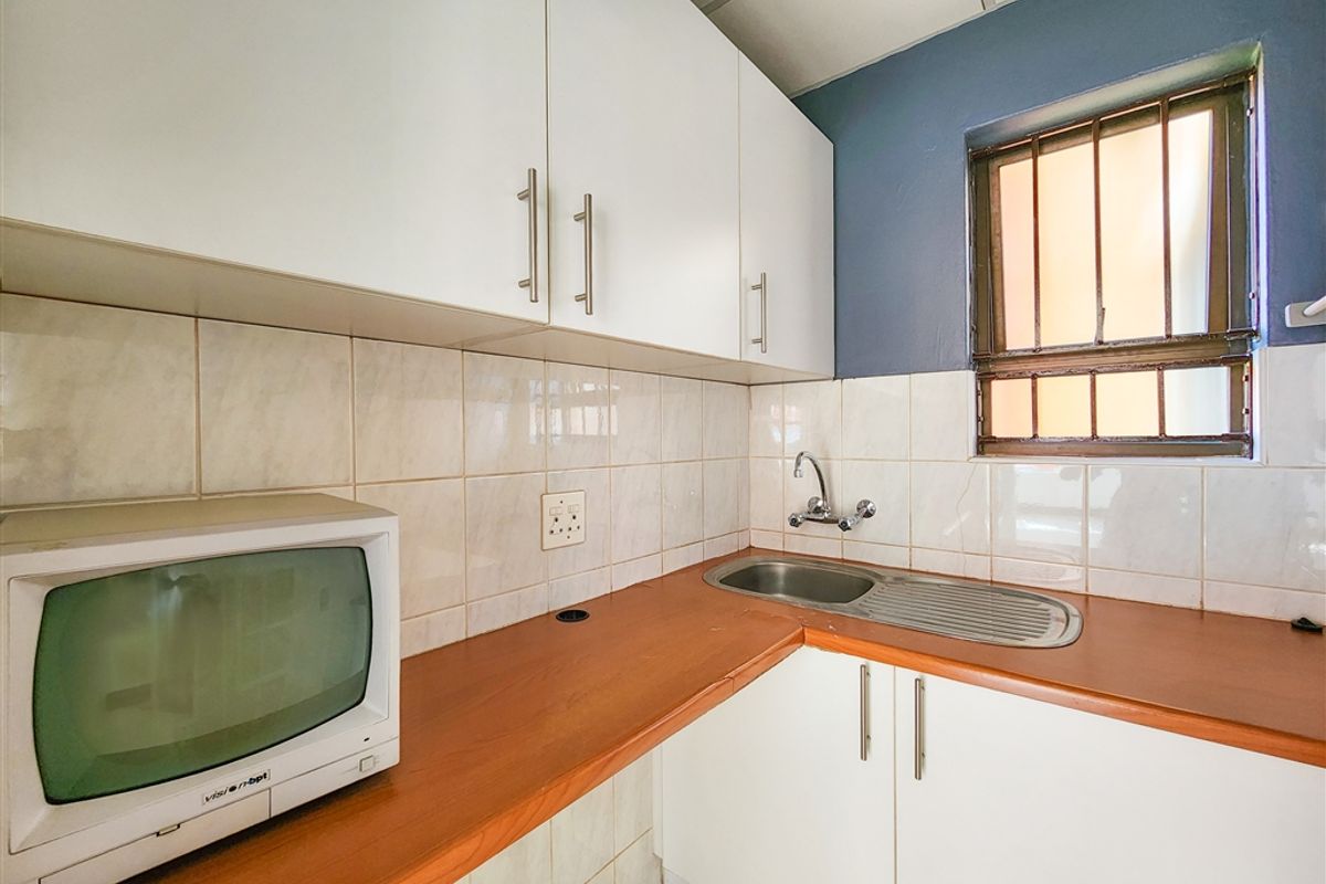 A compact kitchenette featuring white cabinetry, a stainless steel sink, tiled backsplash, and a window with security bars.