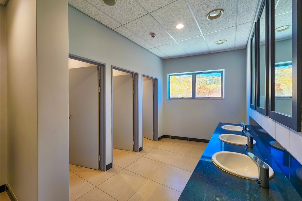 A clean, functional office restroom featuring a long vanity with three integrated sinks, tiled flooring, and multiple private cubicle stalls.
