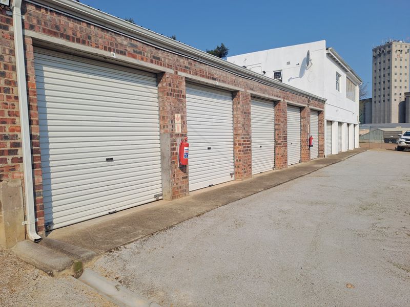 A row of industrial storage units featuring white roller shutter doors, brick construction, and external fire safety equipment on a concrete driveway.