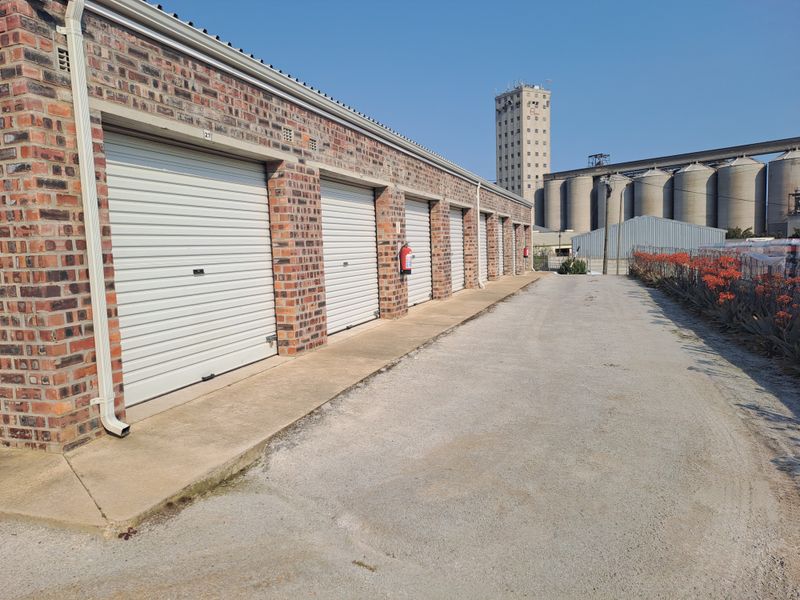 A row of industrial storage units featuring brick construction and secure roller shutter doors, situated on a concrete driveway.