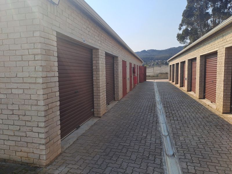 A row of industrial storage garages featuring brick construction, roller shutter doors, and a central paved driveway with integrated drainage.