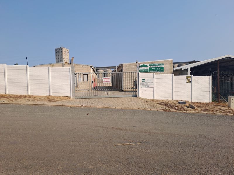 Front entrance view of an industrial storage facility featuring a secure gated access point, paved driveway, and perimeter security fencing.