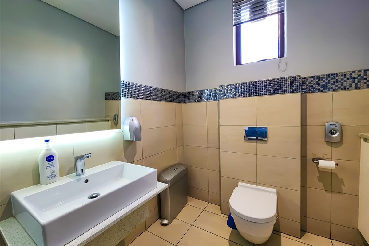 A modern office restroom featuring a wall-mounted basin, a concealed cistern toilet, tiled walls with a decorative mosaic border, and a window with blinds.