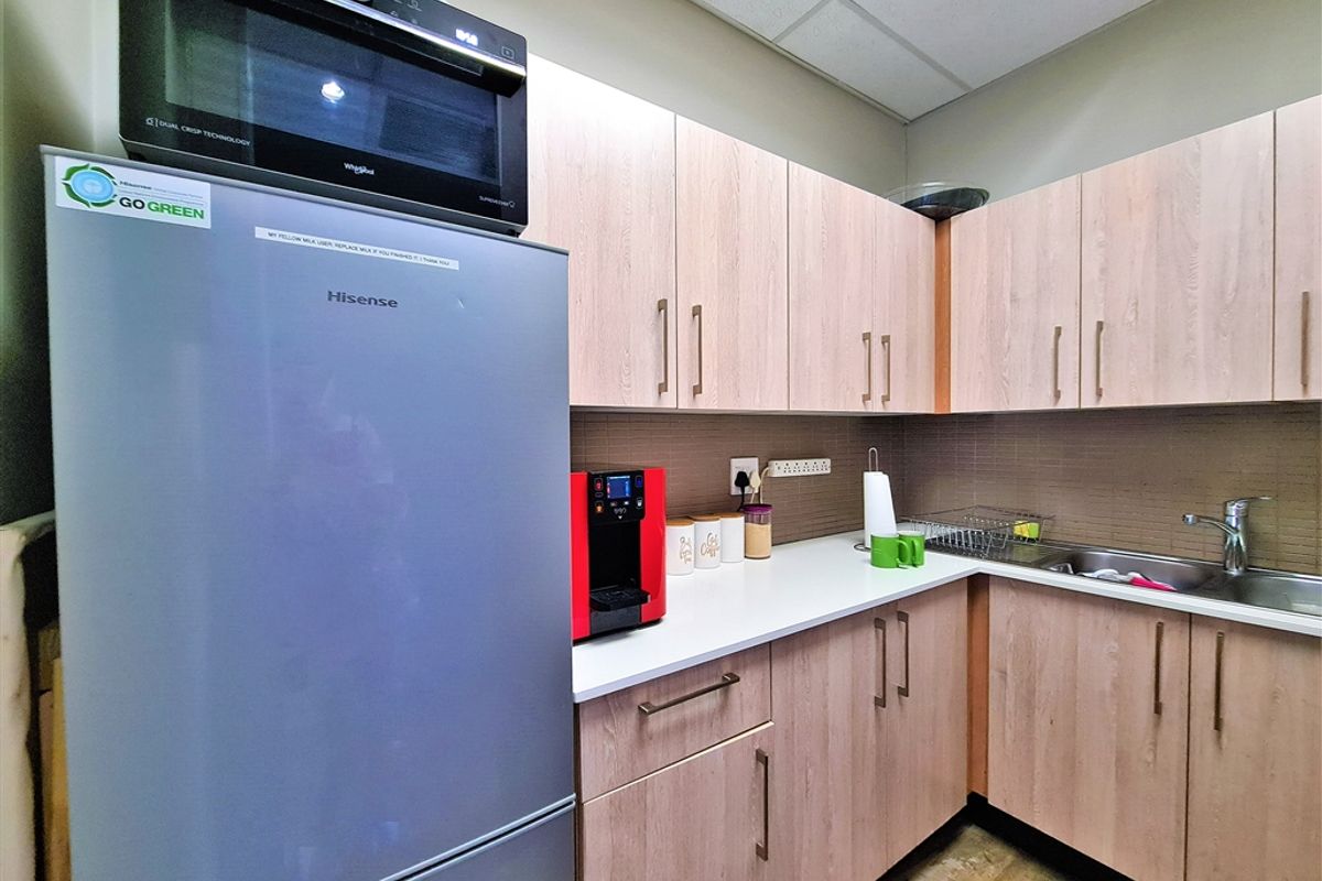 A functional office kitchenette featuring light wood-grain cabinetry, a stainless steel sink, and a white countertop.