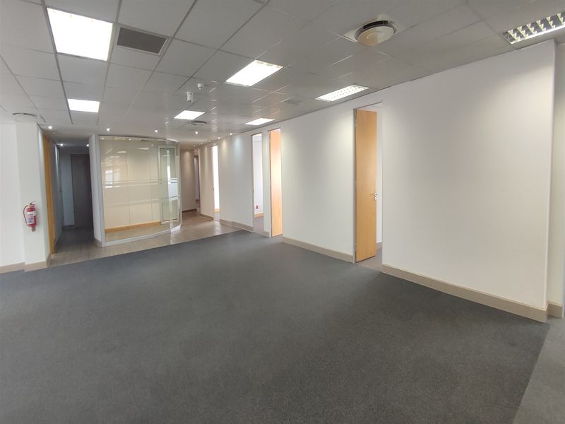 A spacious, empty office area featuring a suspended ceiling with recessed lighting, glass partitioning, and neutral carpet flooring.