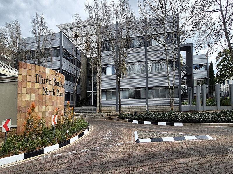 Front view of the Illovo Boulevard North Piazza office complex, featuring a paved driveway, stone signage wall, and modern multi-story office building facade.